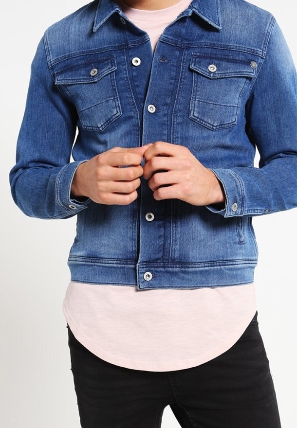 Chaqueta de mezclilla en azul medio con dos bolsillos en el pecho, cierres de botones y costuras en contraste. Llevada sobre una camisa rosa claro con dobladillo curvado.