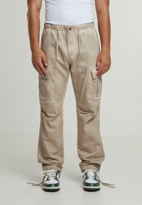 Beige cargo-housut joustovyötäreunalla, joissa on kaksi suurta sivutaskua ja tekstuurikangas. Yhdistetty mustiin ja valkoisiin lenkkareihin.