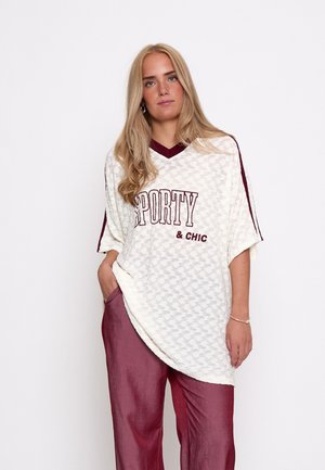 Woman with long blonde hair wears a loose cream top reading "SPORTY & CHIC" and maroon pants, standing against a plain white background.