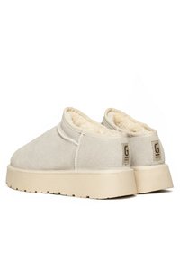 Zapatos de slip-on beige claro con upper de ante texturizado, forro de pelo sintético y una suela de goma gruesa beige. Presenta una etiqueta con el logo en el talón.