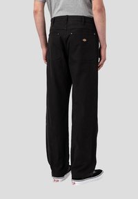 Svarta vida byxor i mjukt tyg, med bakfickor och ett märkesmärke. Kombinerat med svarta skor, visas från baksidan.