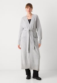 Kvinde iført lang, lysegrå cardigan med bælte, sorte bukser, sorte sko og sølv øreringe, som står mod en ensfarvet hvid baggrund.
