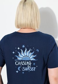 Marine t-shirt met een grafische print van een zon en sterren, met de tekst "Chasing Sunset" in lichtblauw. Korte mouwen en zachte stof.