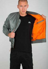 Sivi bomber jakna s narančastom podstavom, s patentnim zatvaračem sprijeda, rebrastim manžetama i rubom, nosi se preko crne majice, ističući elegantan dizajn.