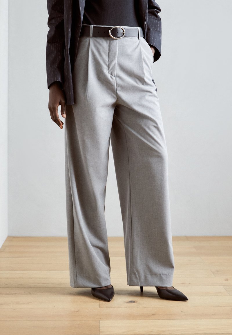 Un pantalon large gris clair avec des plis sur le devant, taille haute et poches latérales, associé à une ceinture noire et des escarpins à bout pointu.