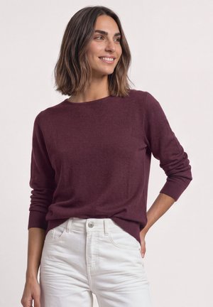 Femme aux cheveux bruns mi-longs souriante, portant un pull bordeaux à manches longues et un pantalon blanc taille haute, debout avec une main sur la hanche.