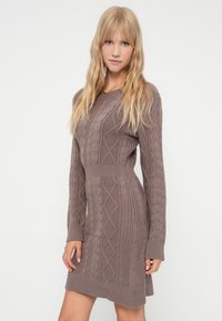 Rochie tricotată în nuanță taupe, cu modele din tricot voluminos, mâneci lungi și talie ajustată. Prezintă manșete și tiv cu bordură rib. 