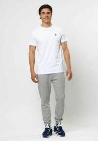 Vit kortärmad t-shirt med en liten svart emblem, som bärs med grå svettbyxor och marinblå sneakers med vita och röda detaljer.