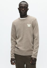 Beige sweater med rund hals og ribbede ærmekanter. Lysefarvet "W"-logo på venstre bryst. Går sammen med khakifarvede bukser. Glat tekstur.