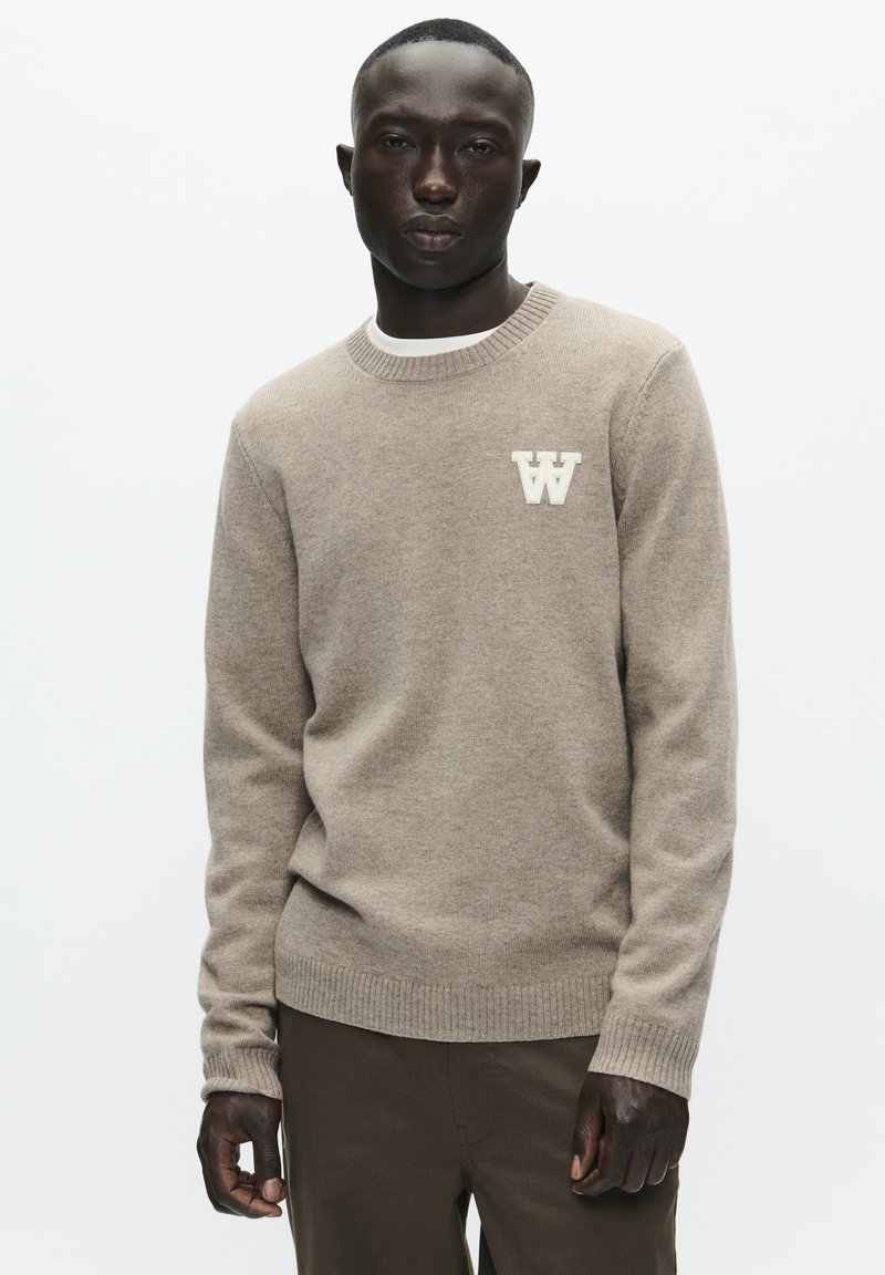 Beige sweater med rund hals og ribbede ærmekanter. Lysefarvet "W"-logo på venstre bryst. Går sammen med khakifarvede bukser. Glat tekstur.