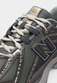 Γκρι και καφέ αθλητικό παπούτσι New Balance που δείχνει το πλέγμα στο πάνω μέρος, το λογότυπο "N Lock" και λευκά και μαύρα κορδόνια περασμένα από τις τρύπες.