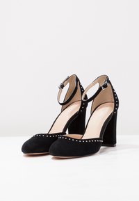 Svarta mocka högklackade skor med rund tå, ankelrem och silvernitar längs kanten. Blockklackdesign.