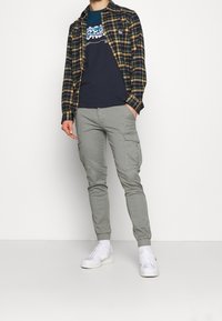 Homme portant un pantalon de jogging cargo gris, des baskets blanches, un t-shirt graphique bleu marine et une chemise à carreaux noire et jaune, se tenant devant un fond blanc.