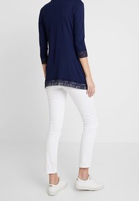 Mujer con una blusa de manga larga azul marino con encaje y pantalones blancos ajustados, de pie contra un fondo claro y uniforme.