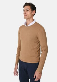 Uomo che indossa un maglione crema a girocollo sopra una camicia bianca con colletto e pantaloni blu scuro, in piedi contro uno sfondo chiaro e semplice.