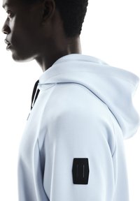 Light blue hoodie with a soft texture, featuring a dark logo patch on the left shoulder. Hood is adjustable with black drawstrings.