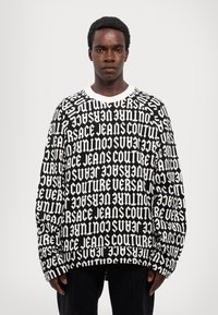 Černá mikina s velkým bílým potiskem „Versace Jeans Couture“ v různých orientacích. Klasický kulatý výstřih, volný střih.