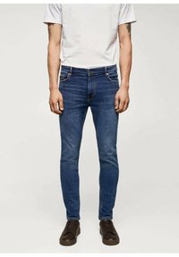 Pánské slim-fit džíny v tmavě modrém denimu. Mají tradiční design s pěti kapsami a zapínání na knoflík. Nošeny s šedým tričkem a tmavými botami.