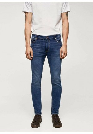 Jeans de corte slim para hombre en denim azul oscuro. Presentan un diseño clásico de cinco bolsillos y cierre de botón. Combinados con una camiseta gris y zapatos oscuros.