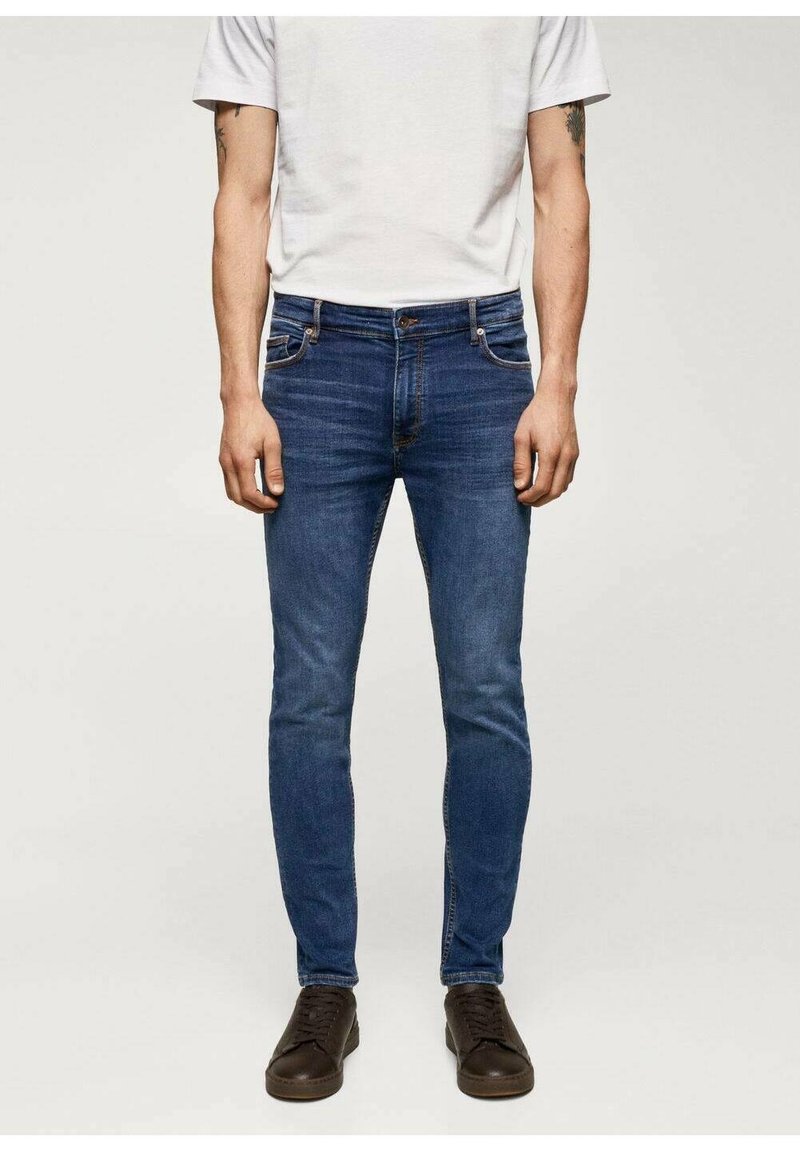 Pánské slim-fit džíny v tmavě modrém denimu. Mají tradiční design s pěti kapsami a zapínání na knoflík. Nošeny s šedým tričkem a tmavými botami.