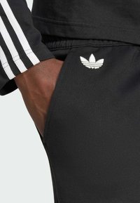 Pantaloni sportivi neri in misto cotone con vita elastica. Presenta un design a tre strisce bianche sulla maniche e un logo ricamato vicino alla tasca.