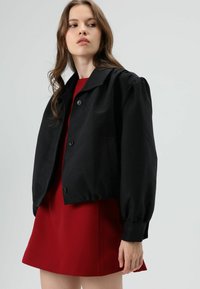 Zwart cropped jack met een kraag, grote opgeblazen mouwen en een knoopsluiting aan de voorkant, gecombineerd met een korte rode jurk. Glad oppervlak.