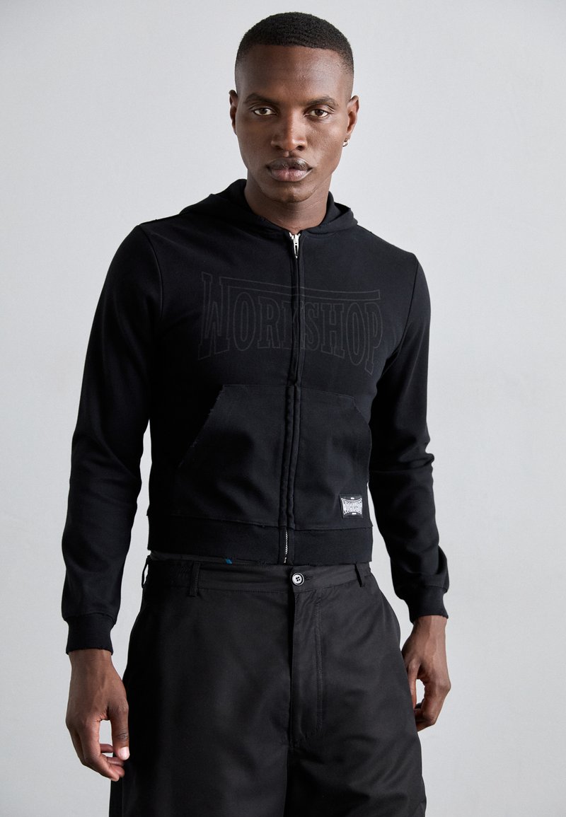 Black zip-up hoodie with a large "WORKSHOP" print on the front, kangaroo pocket, and fitted cuffs. Paired with black trousers.