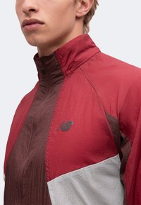 Gros plan d'une personne portant une veste zippée rouge, marron et blanche avec un col montant et le logo New Balance sur la poitrine.