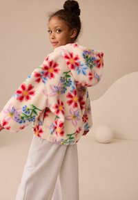 Giacca in pile bianco morbido con motivi floreali colorati, dotata di cappuccio, vestibilità ampia e polsini elastici. Abbinata a pantaloni della tuta grigi chiari.