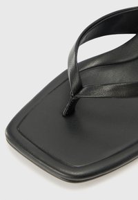 Tongs en cuir noir avec un orteil carré, présentant une texture lisse et de fines lanières. La semelle est plate avec un léger éclat.