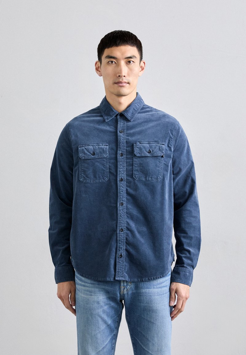 Camisa de manga larga azul oscuro hecha de una tela suave, con un cuello abotonado, dos bolsillos en el pecho y un dobladillo recto.