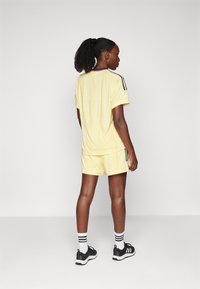 Žltý športový úbor obsahujúci tričko s krátkym rukávom a šortky, s čiernymi pruhmi na bokoch. Nosí sa s čiernymi teniskami a pruhovanými ponožkami.