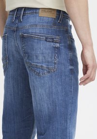 Detalle de un bolsillo trasero de unos jeans de mezclilla azul con una etiqueta de marca de cuero y una pequeña etiqueta de texto, usados por una persona con una camisa blanca.