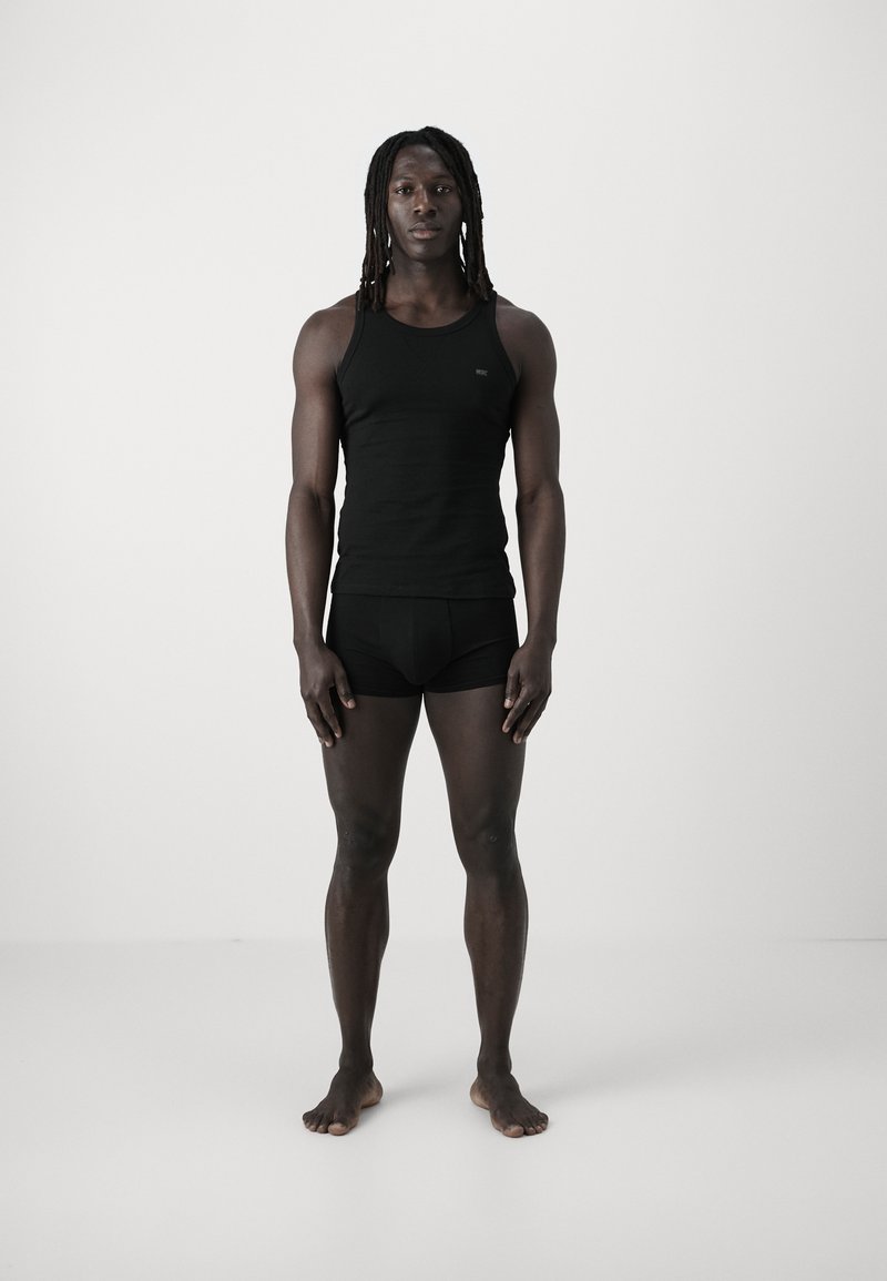 Black sleeveless tank top and matching briefs of soft material, fitted design, worn by a model standing barefoot on a light background.