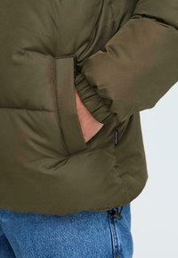 Giacca puffer verde oliva con collarino arrotondato, polsini elasticizzati e una tasca laterale. La texture appare liscia e riflettente, mettendo in evidenza le sezioni gonfie.