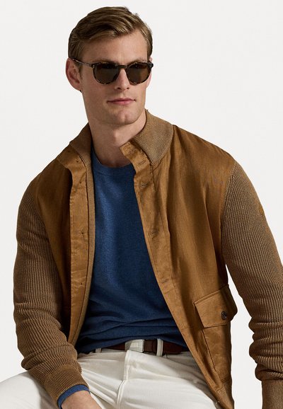 Bruine jas met gestructureerde mouwen, gedragen over een blauwe trui, gecombineerd met witte broek. Zonnebril heeft een donker frame en getinte lenzen.