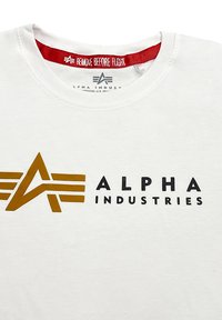 Bílý bavlněný tričko s nápisem "Alpha Industries" v zlaté a černé barvě a červenou etiketou "Remove Before Flight" uvnitř límce.