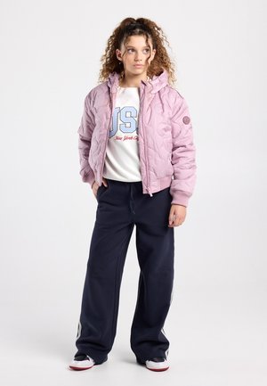 Jonge vrouw met krullend haar, gekleed in een roze gewatteerde jas, een witte sweater met grafische print, marineblauwe wijde broek en zwart-witte sneakers, staand op een witte achtergrond.