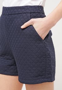 Námorní šortky s diamantovým vzorem, s bočními kapsami a hladkým pasem, zobrazené z boku s rukou v kapse.