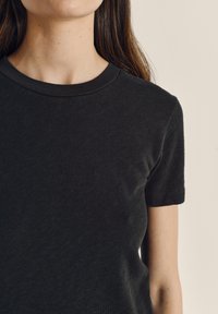 Jaunas žmogus dėvi paprastą juodą trumpomis rankovėmis marškinėlį, matosi kaklas, petys ir viršutinė krūtinės dalis ant neutralaus fono.