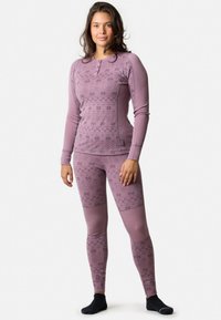 Ung kvinde står i en mauve-mønstret bluse med lange ærmer og matchende leggings med sorte ankelstrømper på en hvid baggrund.