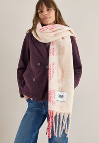 Beige tørklæde med rød og orange mønster, frynser langs kanten, parret med en mørk lilla dobbeltradet jakke og blå jeans. Mærkets label synlig.