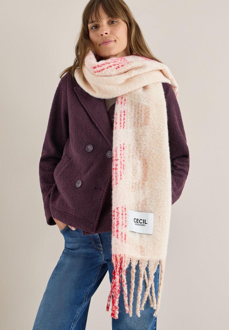 Sciarpa beige con motivo rosso e arancione, con frange ai bordi, abbinata a un giaccone doppiopetto viola scuro e jeans blu. Etichetta di marca visibile.