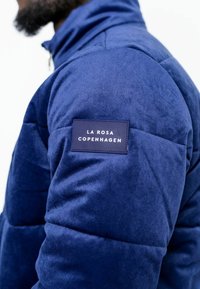 Mörkbl å vadderad jacka i mjuk kordfluga med kviltad textur, med en fyrkantig etikett på vänster axel som säger "LA ROSA COPENHAGEN."