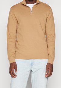 Pullover a quarter-zip marrone chiaro in tessuto testurizzato con colletto e maniche lunghe, abbinato a jeans azzurri.