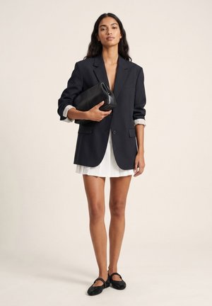 Kvinde i sort oversized blazer over hvid kjole holder sort clutch taske, iført sorte ballerinaer, står mod ensfarvet baggrund.