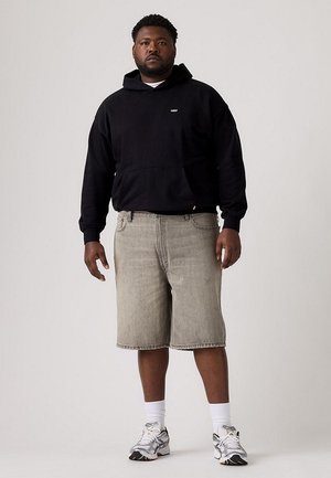 Un hombre posa con una sudadera negra con capucha, pantalones cortos vaqueros de color gris claro, calcetines blancos y zapatillas plateadas, sobre un fondo de estudio sencillo.