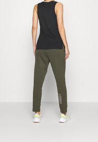 Schwarzes ärmelloses Oberteil kombiniert mit olivgrünen Jogginghosen, die einen elastischen Bund und ein weißes Logo haben. Abgerundet wird das Outfit mit weißen Sneakern.