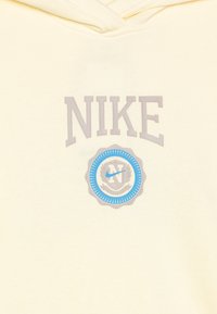 Dzeltena kapucīna jaka ar pelēku "NIKE" tekstu un zilu apaļu logotipu ar stilizētu "N" un lauru akcentiem priekšpusē. Mīksta tekstūra.