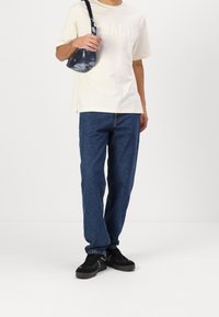 Vit t-skjorta med kort ärm och "COACH"-logotyp, blå denimjeans, svarta sneakers och en liten mönstrad axelväska. Avslappnad outfit.